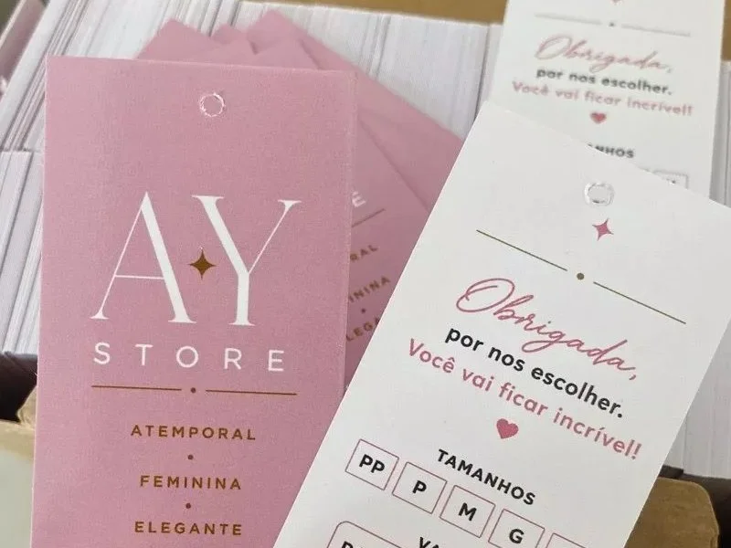 Tags rosa da AY Store com frente e verso, em caixa recém-aberta