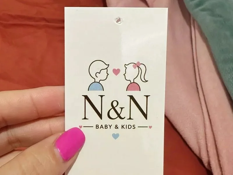 Tag branca da N&N Baby & Kids segurada sobre roupas de bebê